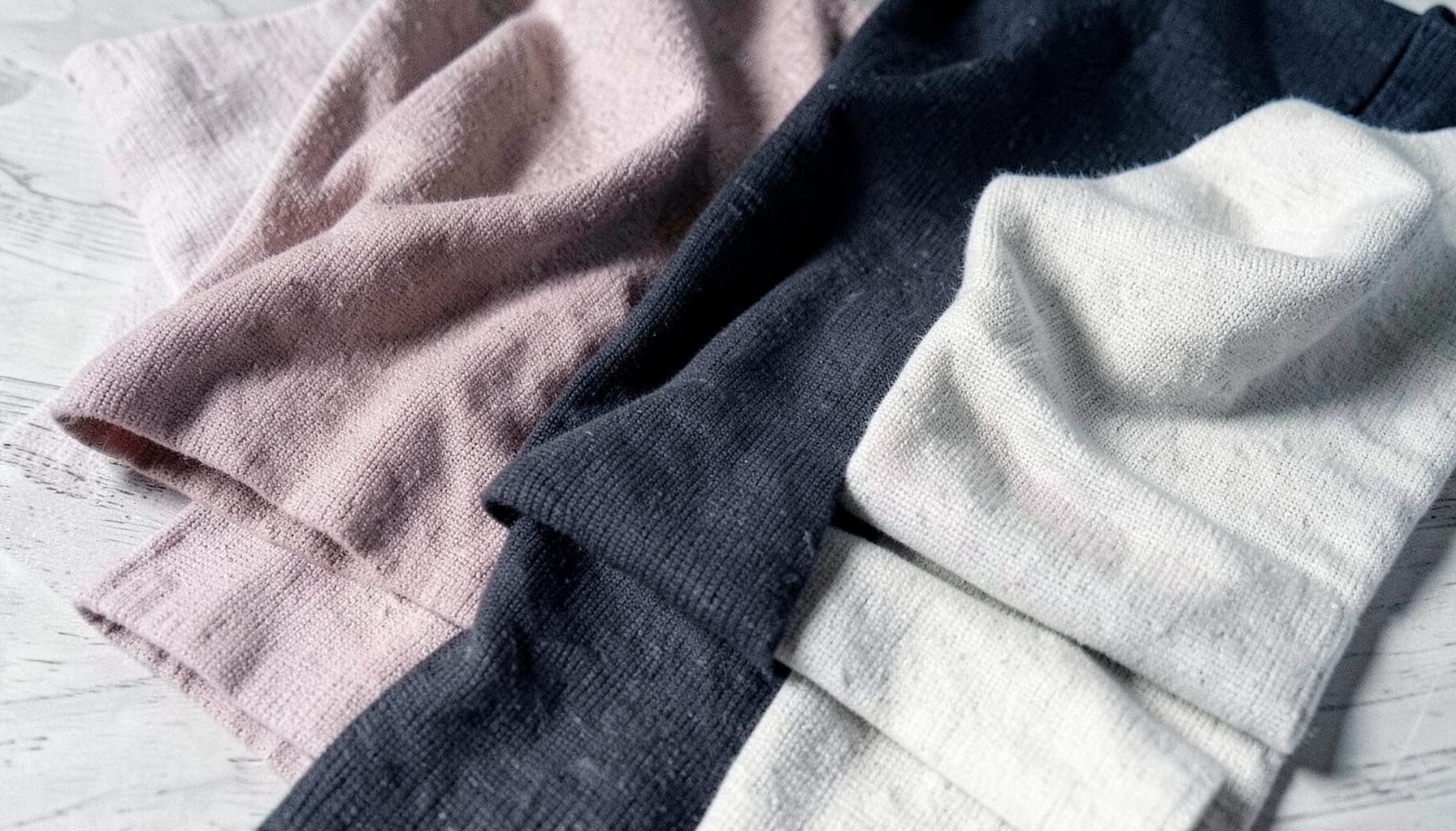 Soft folded knit fabrics in muted blush, charcoal, and cream tones arranged on a light wood surface