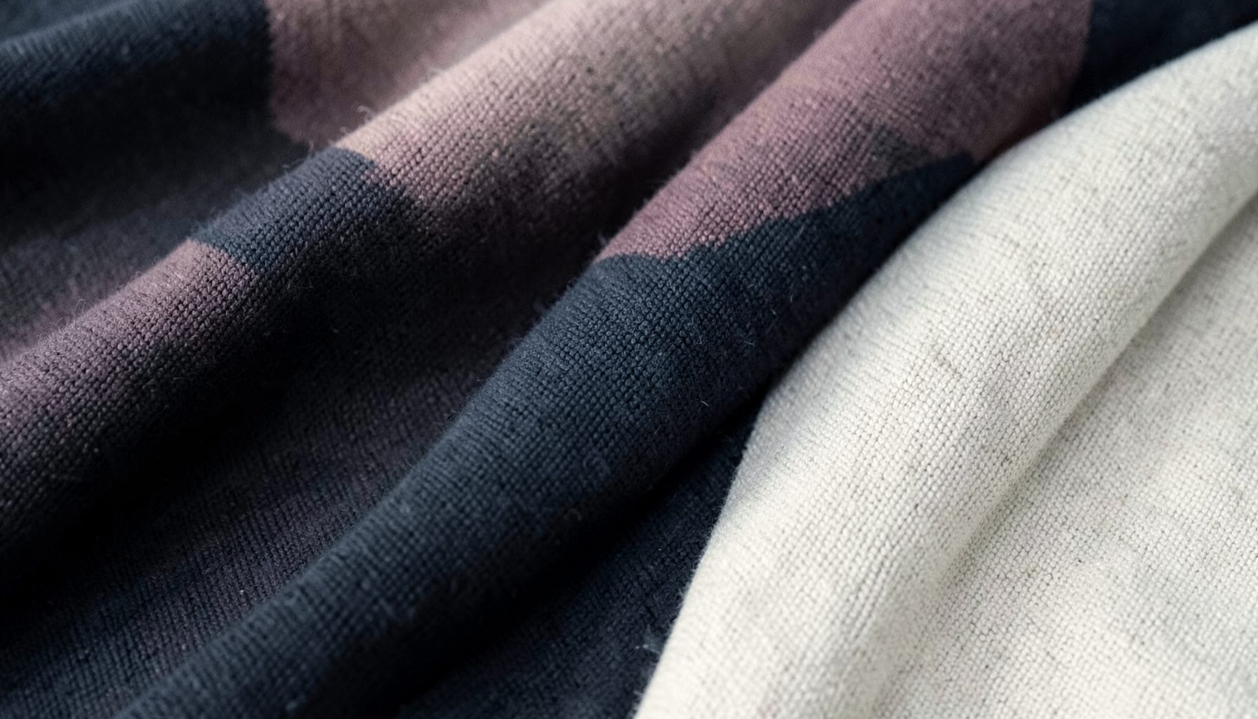 Soft folded knit fabrics in muted blush, charcoal, and cream tones, photographed as a warm duotone with gentle shadows