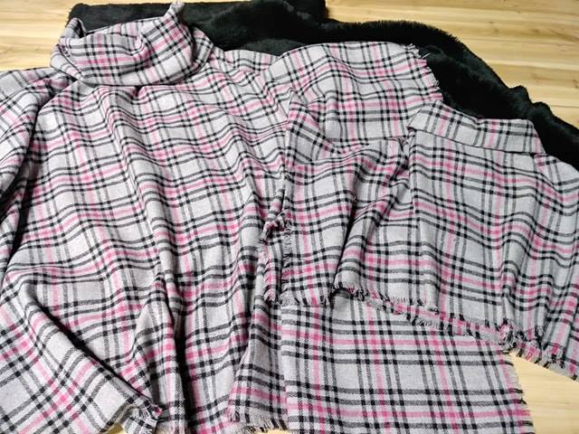 Flat lay of grey, pink, and black plaid flannel fabric alongside black faux fur material on a wooden floor; soft, cozy craft aesthetic