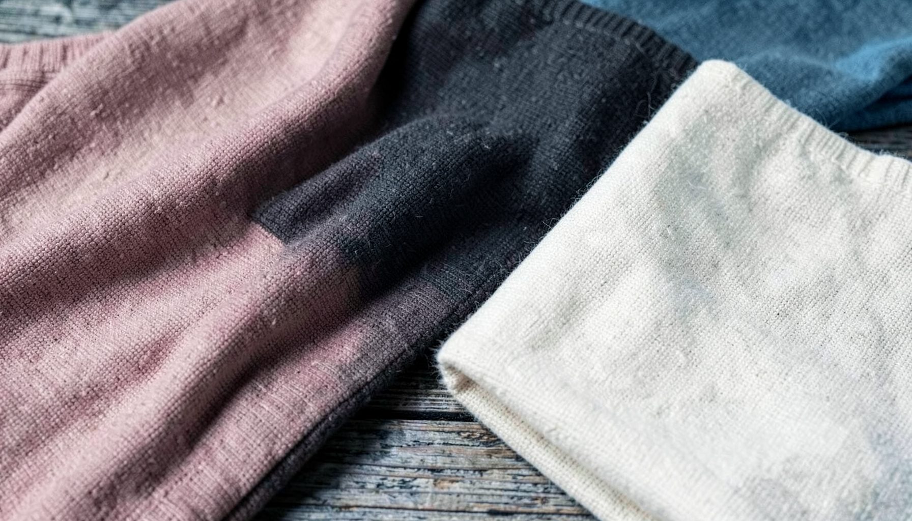 Soft folded knit fabrics in muted blush, charcoal, and cream tones arranged on a rustic wooden surface, warm and inviting textile texture.