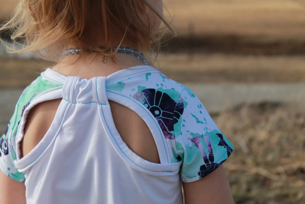 Close-up of a child's back wearing a white and teal patterned top with a cutout design, outdoor blurred background