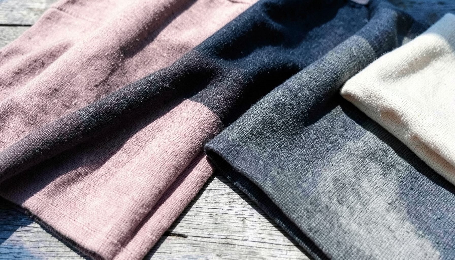 Soft folded knit fabrics in muted blush, charcoal, and cream tones arranged across a sunlit wooden surface